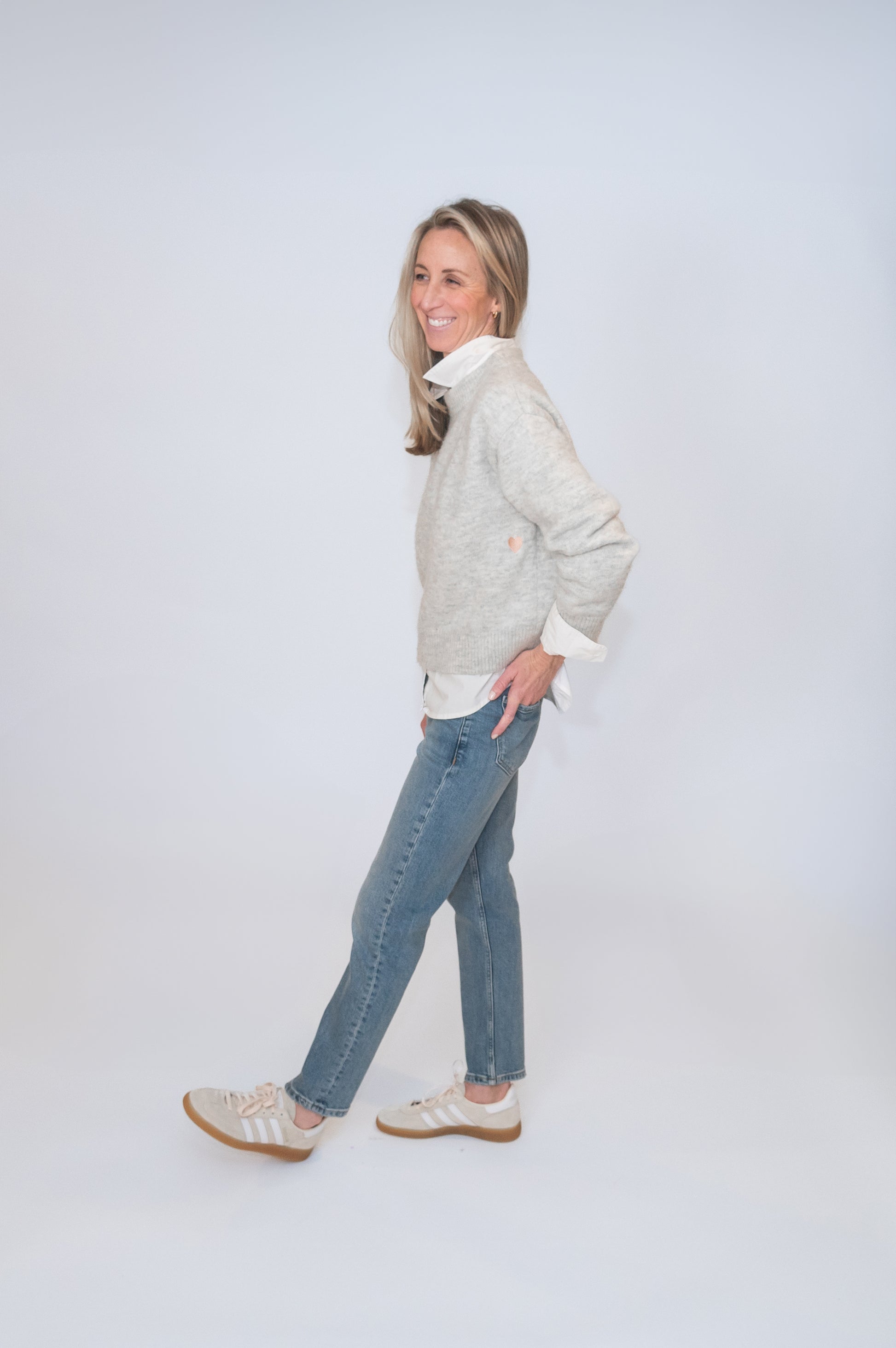 In studio photography of woman wearing a gray cozy sweater with white collared shirt underneath and jeans. She's smiling.