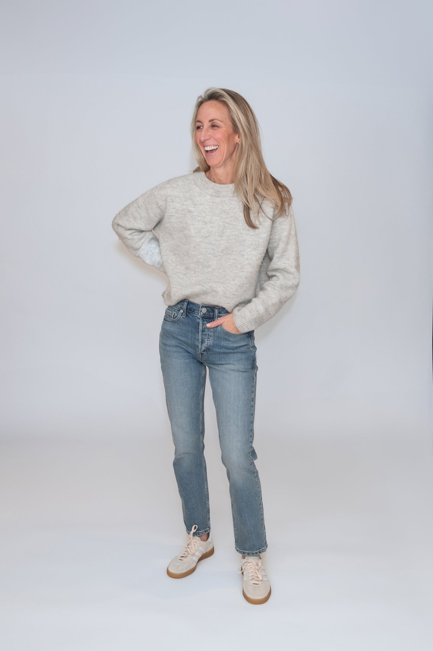 In studio photography of smiling standing woman wearing a gray cozy sweater and jeans.  